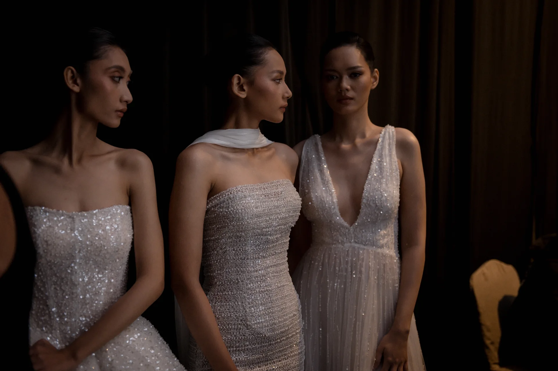 Trio of models backstage in glittering silver sequined evening gowns in ambient warm lighting