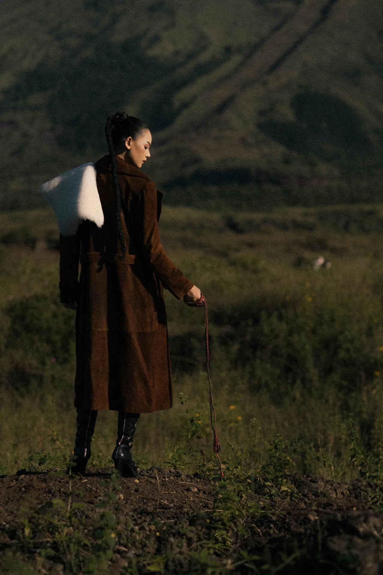 Model in brown suede trench coat with white shearling collar holding walking stick overlooking savanna hills