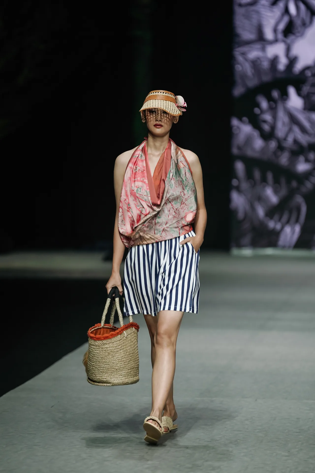 Model walking runway in straw hat, printed halter top, striped shorts and woven basket tote