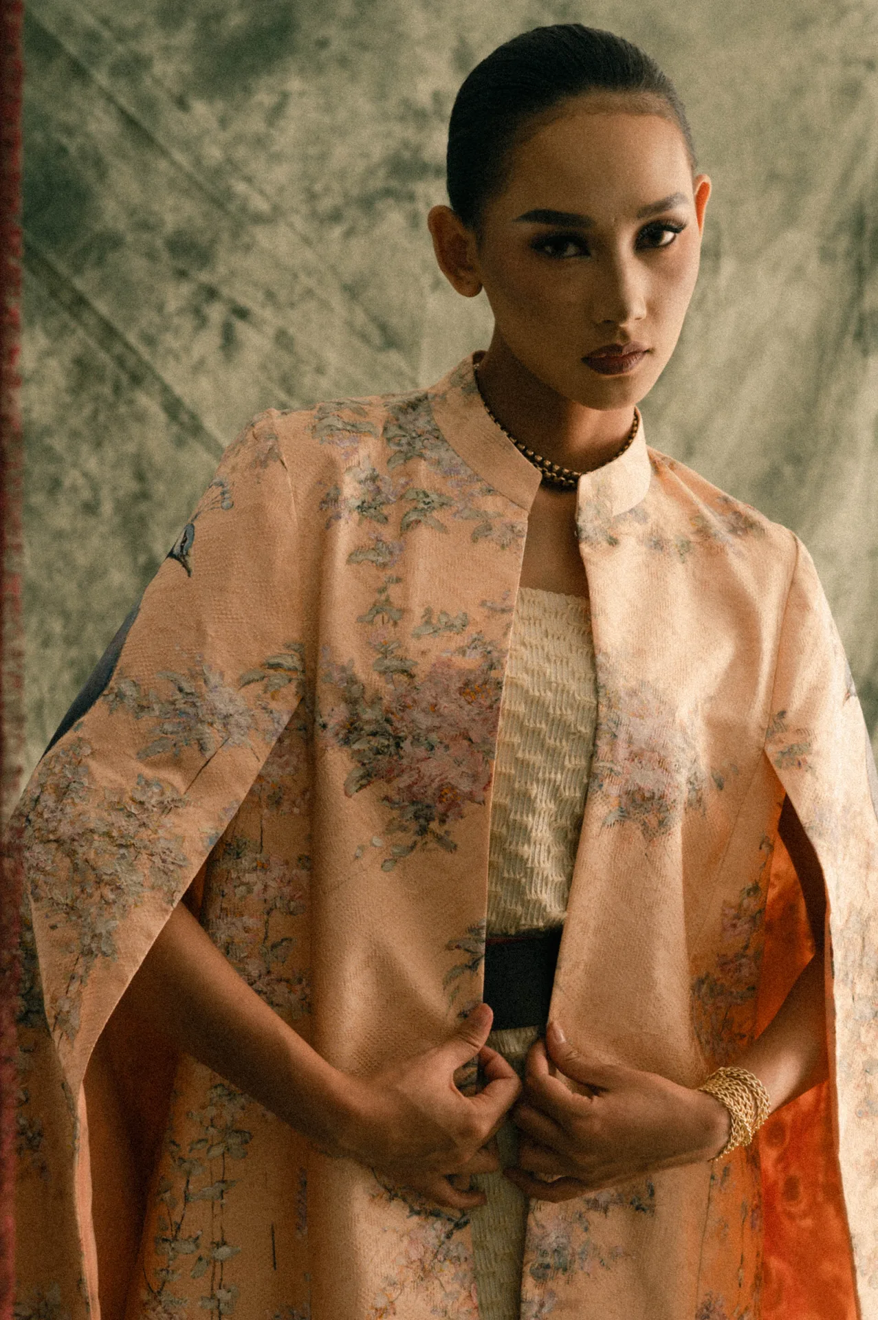 Gina in a peach embroidered cape and textured dress against an olive backdrop